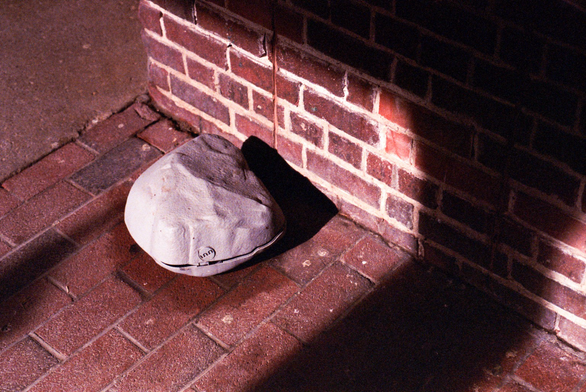 A grey plastic form sits, cracked, on a brick sidewalk catching the sun beside a brick wall.