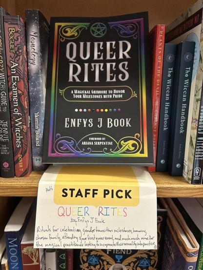 The book "Queer Rites: A Magickal Grimoire to Honor Your Milestones with Pride" on the shelf at a bookstore, with a "Staff Pick" tag beneath it, with a handwritten note saying "Rituals for celebrating gender transition milestones, honoring chosen family, attending your first queer event, and much much more for the magical practitioner looking to incorporate their sexuality into practice."
