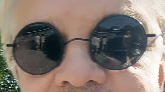 Das Bild zeigt eine Nahaufnahme eines Menschen, der eine runde, schwarze Sonnenbrille trägt. Die Brille reflektiert eine Szene mit Menschen in einem Freibad. Das Gesicht ist nicht vollständig sichtbar, nur der Nasenrücken und Teile der Stirn sind erkennbar. Die Person hat helle Haare.