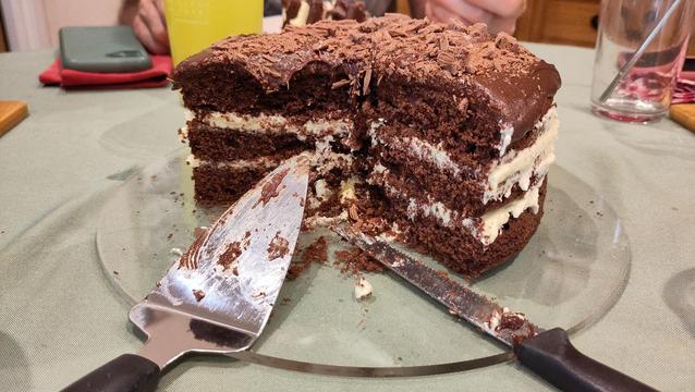 Three layer chocolate cake with creamy icing in between the layers and chocolate icing on top, topped with broken up flake, a large segment has been cut from the cake and a knife and cake slice are lying in the gap.