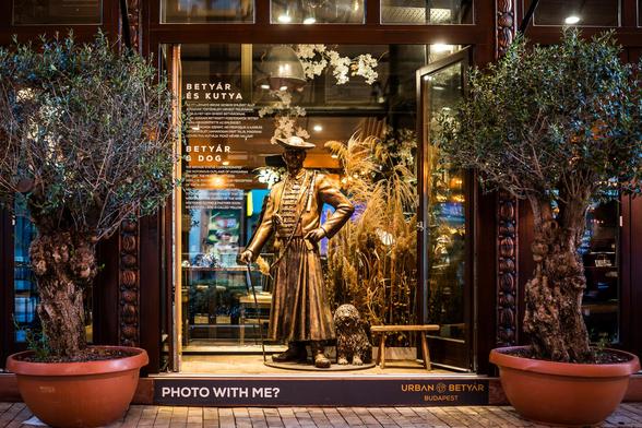 Das Bild zeigt die Schaufensterfront eines Geschäfts mit einer aufwendig gestalteten Installation: Im Zentrum steht eine Bronzestatue eines Mannes in traditioneller ungarischer Kleidung, mit einem Gehstock in der Hand und einem Hund – einem Puli – zu seinen Füßen. Links und rechts der Szene stehen zwei große Kübelpflanzen, die das Ensemble einrahmen. Hinter der Glasfront erkennt man den Schriftzug „URBAN BETYÁR BUDAPEST“ sowie erklärenden Text zu der Darstellung.

Der Text beleuchtet die Geschichte der sogenannten Betyárok – ungarische Straßenräuber aus dem 18. und 19. Jahrhundert, die vom Volk als Freiheitskämpfer und Helden angesehen wurden. Ihre Kleidung stammt aus der ungarischen Volkskultur, besonders auffällig ist der „szűr“ – ein Hirtenmantel. Der Hund an ihrer Seite, der Puli, ist ebenfalls ein Symbol des ungarischen Hirtenlebens.

Unten auf der Glasfront lädt ein Schild die Passanten ein: „Photo with me?“ – ein charmantes Detail mit einem Augenzwinkern. 📸