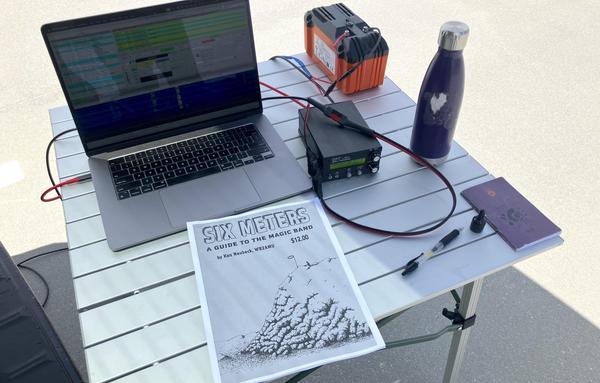 A silver laptop, orange battery, notebook, purple water bottle and small black transceiver sit on a square metal table, along with a print out of some pages from the book "Six Meters: A Guide to the Magic Band" by Ken Neubeck, WB2AMU