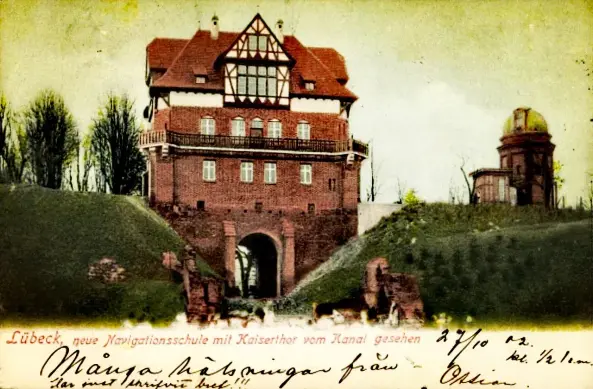 The image appears to be a postcard featuring a historical building in Lübeck, Germany, likely depicting a navigation school. It shows architectural elements typical of the region, including a tower and a bridge. There are annotations and handwriting at the bottom of the postcard, suggesting it may have been sent or addressed to someone. The overall style and coloring suggest it may be an older, possibly hand-colored postcard.

Image Credits: Wikimedia / Anonymous photographer, not identified anywhere; no publisher mentioned either / Public domain
