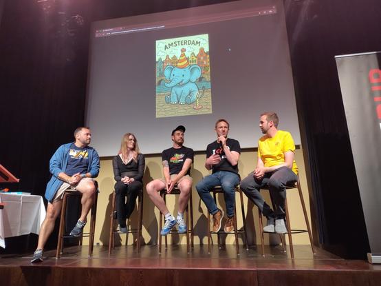 5 people on stage, a slide in the back with an elephant with a party hat and the word Amsterdam on top