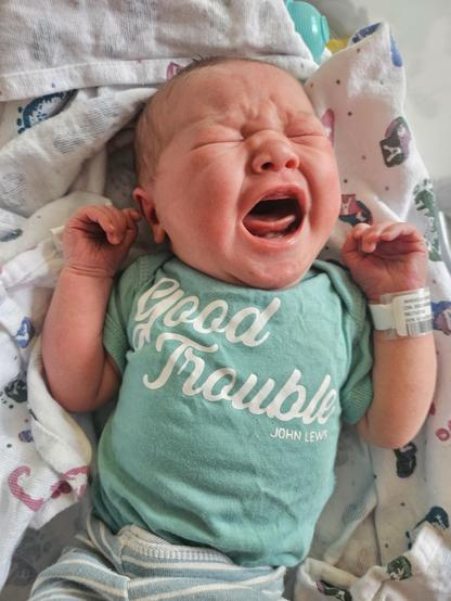 A newborn baby in a hospital setting. The baby is wearing a light green shirt with the text "Good Trouble – John Lewis.” The background features a patterned blanket