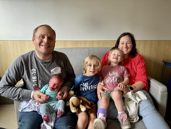 A family of five is seated together on a couch. The father holds a newborn baby, while the mother smiles next to him. Two older children, a boy and a girl, are positioned in front, wearing coordinated shirts that say "big brother" and “big sister”
