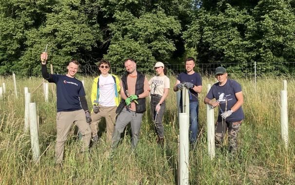 Das Bild zeigt eine Gruppe von sechs Personen, die gemeinsam auf einer Wiese mit hohem Gras arbeiten. Sie stehen zwischen jungen Bäumen, die durch helle Kunststoffhüllen geschützt sind – vermutlich bei einer Pflanz- oder Pflegemaßnahme.