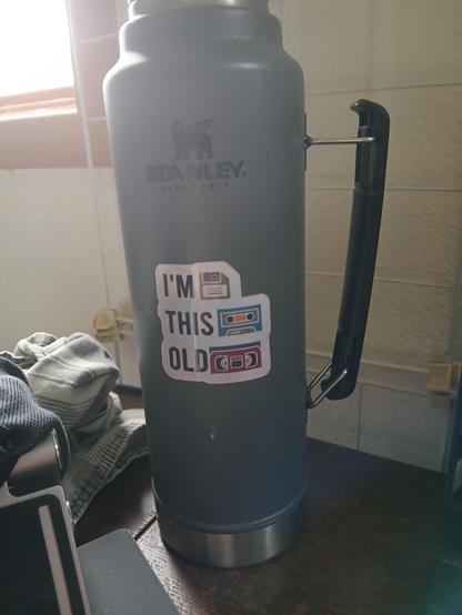 A large Stanley vacuum flask with a sticker on it, and the sticker says...

I'M (image: 3.5" floppy disk)
THIS (image: audiocassette tape)
OLD (image: VHS cassette tape)

The flask is full of strong black tea.
