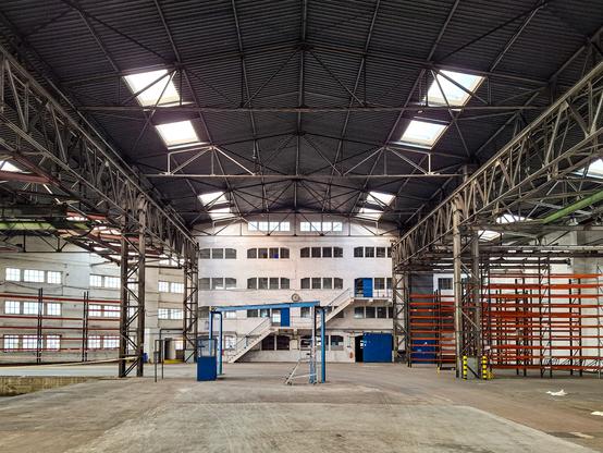 Eine geräumige Industriehalle mit einer hohen, metallenen Decke, die von zahlreichen Oberlichtern durchbrochen wird, füllt das Bild. Die Deckenkonstruktion wird von massiven Stahlträgern gestützt. In der Mitte der Halle steht ein weißes, mehrstöckiges Gebäude mit vielen Fenstern und mehreren blauen Türen und Treppen. Links und rechts des Gebäudes sind weitere Fensterfronten zu sehen. Rechts im Bild befindet sich ein großes Regalgerüst mit orangefarbenen Streben. Der Boden der Halle ist aus Beton. Die Halle wirkt leer und ungenutzt.