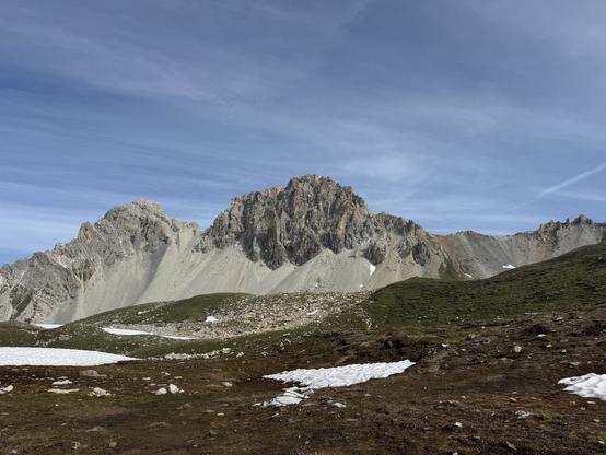 South face of Piz digl Gurschus. Rocky ridge with vast light gray scree fields below. Alpine meadows with small patches of snow in the foreground.