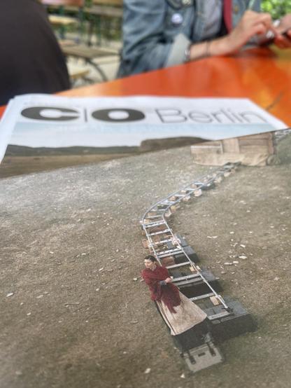 Eine Zeitung liegt auf einem Biergartentisch. Das Titelbild der co-berlin-Zeitung ist ein Bild von Julian Rosenfeldt. Es zeigt eine Kameraschlittenführung inmitten der Wüste. Im Vordergrund eine Schauspielerin im roten Kleid.