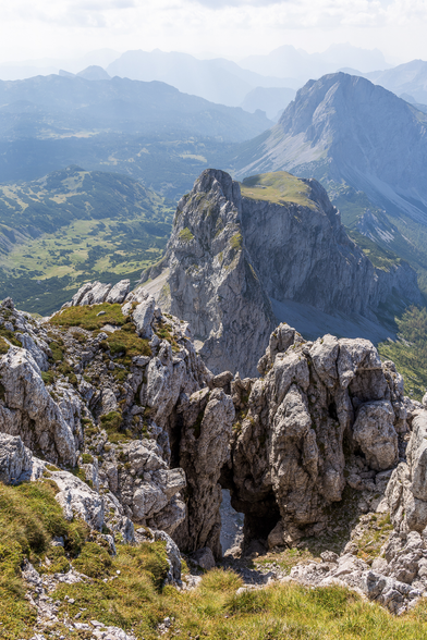 Hochformat einer steiler abfallenden Stelle auf einem Berg. Direkt vorne sind Felsen, mit einer Spalte in der Mitte. Dahinter geht es offensichtlich steil nach unten. Dahinter sieht man einen schroffen alleinstehenden Berg. Wiederum dahinter gibt es noch viele Gebirgszüge in mehreren Layern.