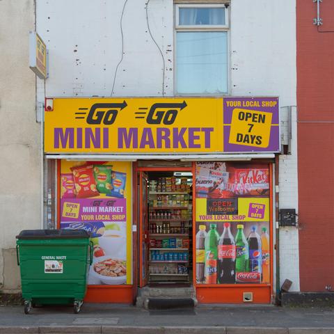 A single terrace house width store called Go Go Mini Market, it's windows covered in product pictures but the open door shows the inside stacked full of items to sell. Outside and to the left is an industrial size green general waste bin.