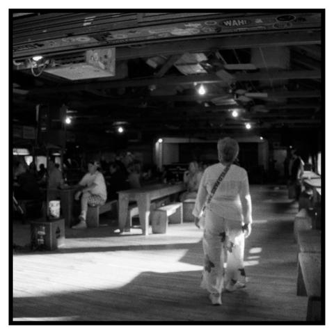 Photo of an older woman, clothed in white walking into Gruene hall in the early summer evening. Light streams in theough windows on the right and to the left there are people sitting and watching a local musician perform on acoustic guitar.