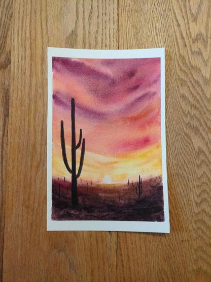 This is a watercolor landscape painting of Saguaro National Park at sunset with the dark silhouettes of the saguaro cacti. This is painting in portrait orientation. The sky starts out purple, orange and pink in big swashes of color with granulation from Schmincke Wild Purple. Closer to the setting sun, the sky shifts to orange and warm yellows. The landmass is orangey brown near the horizon line and then more purpley brown in the foreground.  There is one large cactus with three arms and many small cacti in the background.