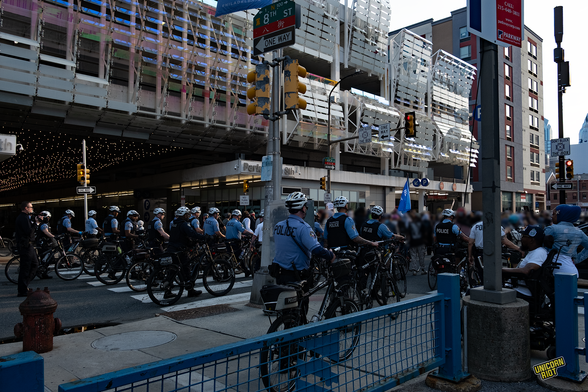 Photo of a street scene in Philadelphia. In the front there are dozens of police officers holding their bicycles. a crowd of protestors in the back. in the background a modernist building