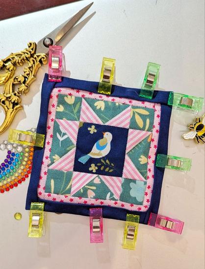 Light grey  surface, on it a small quilt block in dark blue, pink and green. The pattern ist like a star around an inner square (with a bird motif). The dark  blue border is hold with cloth clips. Golden scissors and a needle minder in bee form lie next to the unquilted Quilt block.