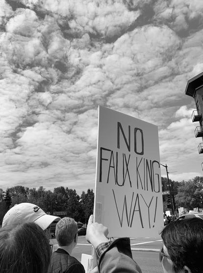Black and white photo of a sign at a protest that says "NO FAUX KING WAY!" amongst some pretty clouds.