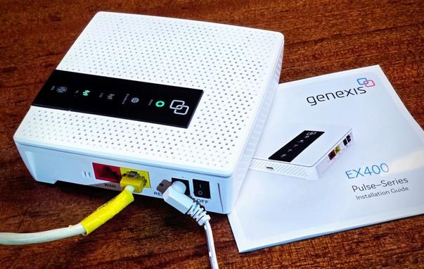 Ein Wlan-Router steht auf einem Holztisch, darunter eein Anleitungs-Büchlein. Der Router ist weis, quadratisch und hat ein schwarzes Bedienpanel mit mehreren LEDs auf der Obeseite, auf der Seite zwei Lanbuchsen rot und gelb, eine Reset-Öffnung, ein DC-Kabel steckt gewinkelt, ein ON/OFF-Schalter.