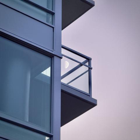 An apartment balcony railing's plexiglass sidewall reflects the moon's reverse image on a clear twilight morning.