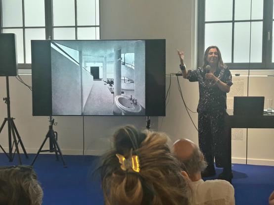 A woman holding a microphone and gesturing while talking next to a large screen. The screen shows a black and white picture of a modernist interior consisting of only concrete, steel and glass, with a large sink prominently pictured next to the entrance.