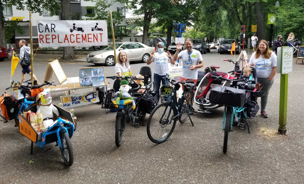 Group of people in BikeLoud t-shirts standing behind their cargo bikes. A table with material and a sign above the table that says "Car Replacement"