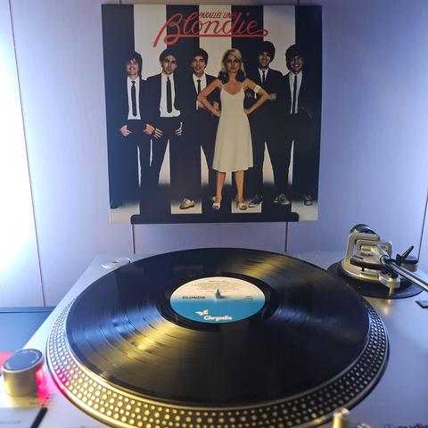 A vinyl record sits on a turntable. Behind the turntable, a vinyl album outer sleeve is displayed. The front cover shows the members of Blondie standing together with black and white vertical stripes in the background. The 5 men are wearing black suits w/ white shirts and black ties. Deborah Harry is standing in front in a white dress and her fists on her hips.