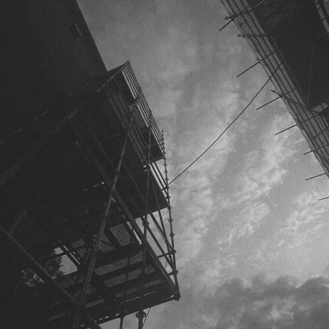 A black-and-white grainy photo of the sky and buildings covered in scaffolding, and a cable or rope hanging between the scaffolds