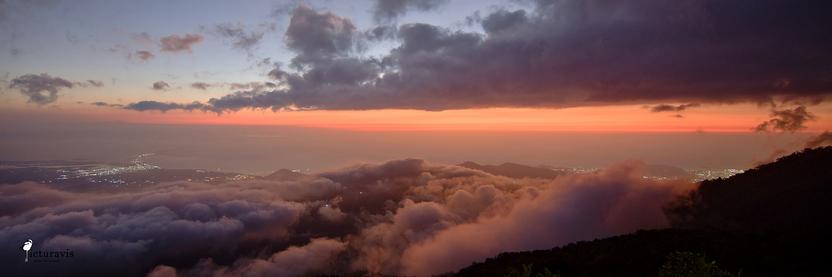 A panorama photo taken from high on a mountain looking towards the Caribbean sea. Most of the view is blocked by cloud lines above and below the photographer, beautifully illuminated by the orange setting sun. Part of the mountain is visible on the lower right. Near the coast are two areas with tiny white spots, where city illumination is visible through gaps in the clouds. The sky is a gradient from blue to reddish orange.