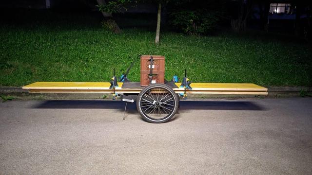Ein Radanhänger ohne Deichsel steht auf Asphalt. Auf dem Anhänger liegen lange gelbe Schalungsplatten die mit Zwingen am Anhänger und mit sich selbst fixiert werden. Die Platten sind 3m lang. Der Anhänger knapp 80cm. Die Platten stehen also vorne und hinten darüber hinaus