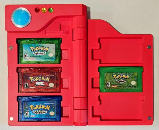 A bright red, Pokedex-shaped, bifold case, open to reveal four Game Boy Advance cartridges. The left side holds three cartridges stacked vertically: a translucent green "Pokemon Emerald Version", a translucent red "Pokemon Ruby Version", and a translucent blue "Pokemon Sapphire Version". The right side holds a single, solid green "Pokemon LeafGreen Version" cartridge with a swirling pattern. The case itself features a circular translucent blue lens and three small colored "indicator lights" (red, yellow, green) on the top left, consistent with its Pokedex design. It appears to be made of 3D-printed plastic.