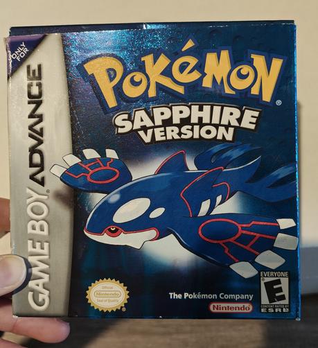 The front of a "Pokemon Sapphire Version" video game box for the Game Boy Advance. The predominantly dark blue box has a metallic finish and features a large illustration of the legendary Pokemon, Kyogre, in the center. Kyogre is depicted as a large, whale-like creature with dark blue skin, white undersides, and striking red markings. The box shows some signs of wear and is being held up for viewing.