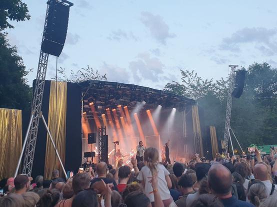 The main stage in the evening on Saturday, lit up by spotlights. It is not dark yet, and the sky above has a light blueish shade. In front of the stage the crowd is standing, with a little girl in a white summer dress sitting on someone's shoulders.
