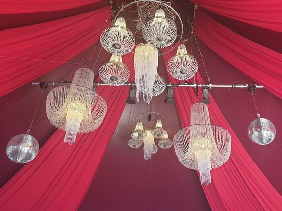 A view of the inner top of a large stage tent in two shades of red, decorated with several silvery and shiny chandeliers and mirror balls.