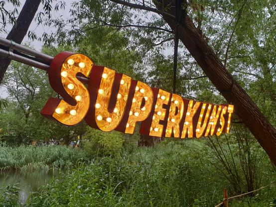 One part of the decoration: a large sign consisting of orange and lit up single letters making up the word "Superkunst" in front of a green nature background.