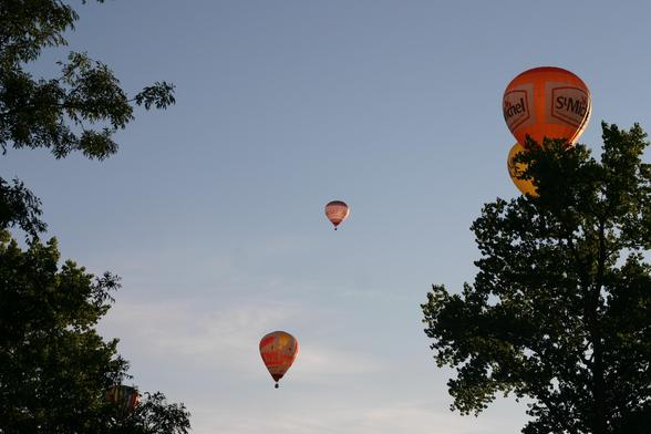 Photo horizontale. Encadrées en bas par des ramures d'arbres, on aperçoit 4 montgolfières, à différentes altitudes, de différentes couleurs dans une lumière de début de soleil couchant.
Coupe d'Europe de montgolfières - Mainfonds - Charente - France - 08/2017