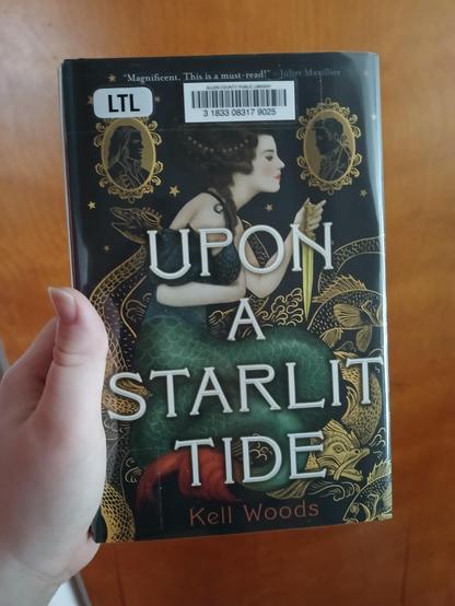 My hand holding up a copy of "Upon A Starlit Tide" by Kell Woods.