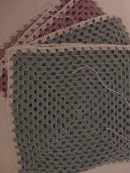 Sechs gehäkelte und noch nicht vernähte Granny Squares, die aufeinander liegen, bestehend aus Filetgarn. Die oberen drei sind mintgrün, mit weißem Rand und die unteren in einem altrosa mit weißen Rand.