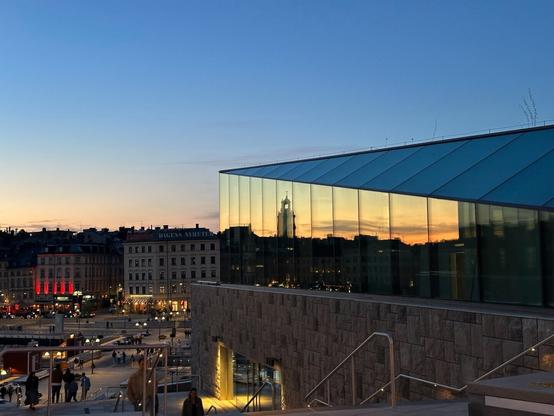 A view down a wide outdoor staircase descending a terrace, on its side a stone-clad building with an angular glass top. Beyond it is a set of bridges crossing water towards an old European city area, with lit-up restaurant/bar-type establishments. The side of the angular glass building reflects a golden sunset and the silhouette of a tower.