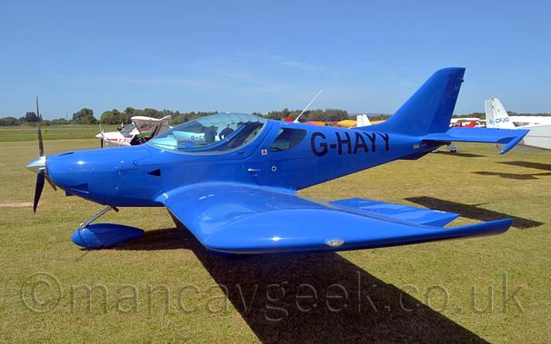 Side view of a single engined light aircraft parked on grass facing to the left.
The plane is a vivid blue, with black registration "G-HAYY"  on the rear fuselage.
A large bubble canopy covers the 2 seat cockpit.
The plane is casting a strong shadow almost straight down on the ground, suggesting it is sunny and the sun is almost overhead.
A collection of light aircraft are parked behind this plane, with trees lining the airfield perimeter in the distance, with bright blue sky filling the rest of the frame.