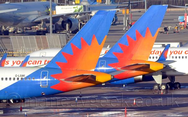 Close-up of the rear fuselage and tail of a pair of near-identical jet airliners parked facing to the left.
The planes have white bodies, with a blue rear fuselage and tail, with a large red and orange sun burst.
The nearest plane has the black registration "G-LSAK" on the upper rear fuselage.
The second plane has some red, fairly indistinct text running along the body.
There is a third plane from the same airline showing most of it's fuselage behind them.
Another white jet airliner is in the background on the left.
Bright sunlight floods the frame, causing reflections all over the place.
