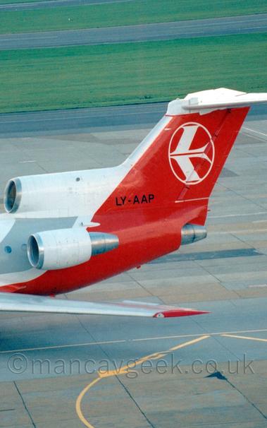 Close-up of the rear fuselage and tail of a 3 engined jet airliner taxiing from right to left.
The plane has a white body with a red belly and a grey stripe covering the passenger cabin windows.
The tail and rear fuselage are red, with a stylised white bird in the white outline of a circle in the middle of the tail.
The white engines are mounted on the top and sides of the rear fuselage , with the centre engine intake being built in to the front of the tail, but exhausting below it.
The black registration "LY-AAP" is at the bottom of he tail.
Grey and black concrete apron fills the foreground, with black taxiways divided by large grassed areas fills the background.