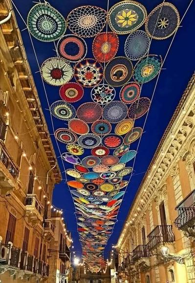 Photo of a street at night looking up between two rows of golden buildings with balconies, to a strip of colourful crocheted circles joined together under a deep blue night sky