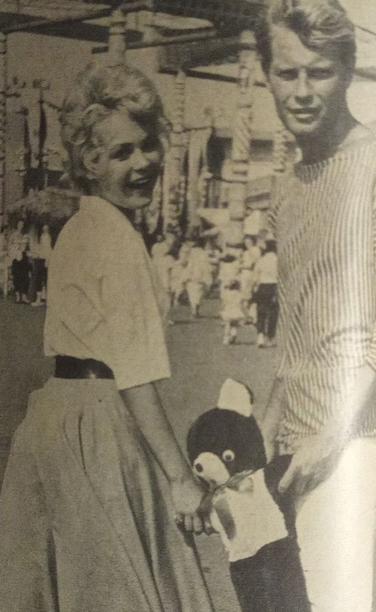 Sandra Dee y pretendiente  sonrientes sostiene un peluche mientras pasea en un lugar público, rodeada de gente.