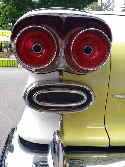Forgive me, I don't recall the make of this vintage car.
But it is looking at the left, rear tail light.
The car is two-tone. Yellow and white. There are two large, round, red, tail lights with a sort of chrome eyebrow that covers both.
The tail lights have a small round black center - which makes it look like eyes.
There is another design element - not for sure why, but it is oval shaped and just below the tail lights, giving this view a 'surprised' or ' oh boy!' look.