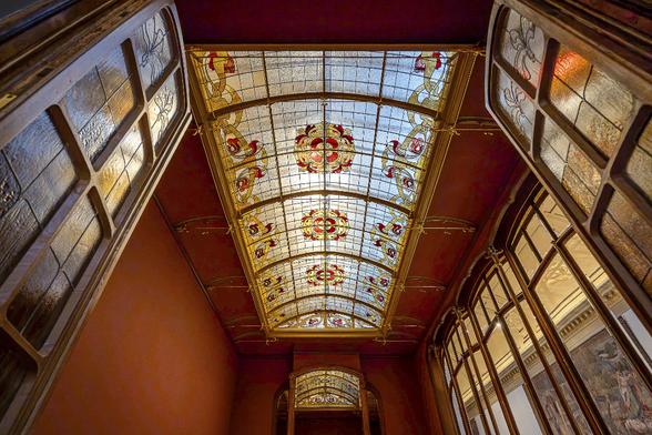 Look up to the Art Nouveau stainglass ceiling ornated with red and yellow flowers of the winter garden of the Cousin House, designed by Victor Horta. This winter garden has just been restored and reassembled and is now available to see at Brussels Art and History Museum since June 2025.