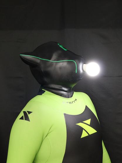 Person wearing a green and black Xterra wetsuit and a black Furrjoi drone mask with glowing green accents, facing sideways with a lit light bulb in their mouth - symbolizing a sudden idea or creative inspiration.