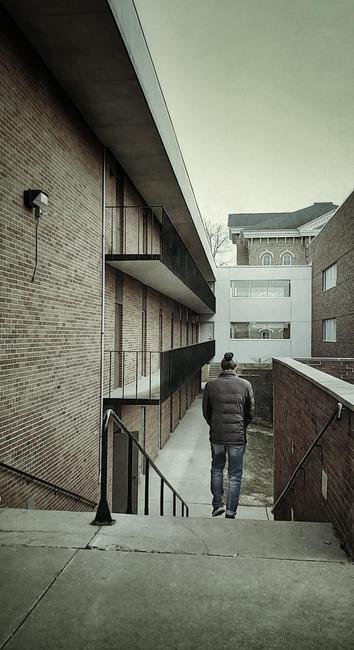 A person walking down a set of steps into a passage way between buildings.
They are seen from behind. Wearing jeans and a coat.
The convergence of balconies, roof lines, and walls all point to the center of the photo.
