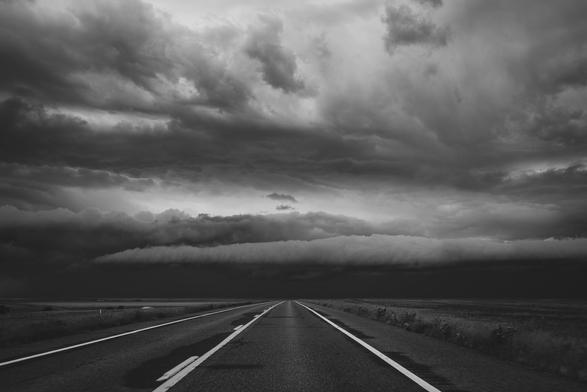 Looking over a country road, toward dark clouds on the horizon. Clouds in front appear to be rolling into a tube.