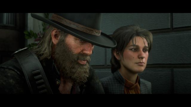 The image is a screenshot from the video game Red Dead Redemption 2. In the foreground, there are two male characters. The character on the left appears to be older with a beard and mustache, wearing a brown cowboy hat and black attire. He is facing away from the camera towards another character who is standing in front of him. This second character has short dark hair and is also facing away from the camera towards the first character. Both characters are set against an urban background with buildings visible in the distance. The image suggests a moment of interaction or conversation between the two characters during gameplay.